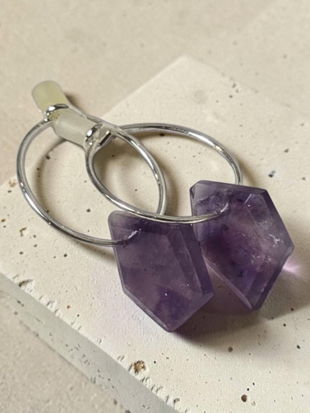 reveriebygemma Lucid Drops amethyst hoop earrings featuring faceted purple gemstones on silver hoops on a textured surface