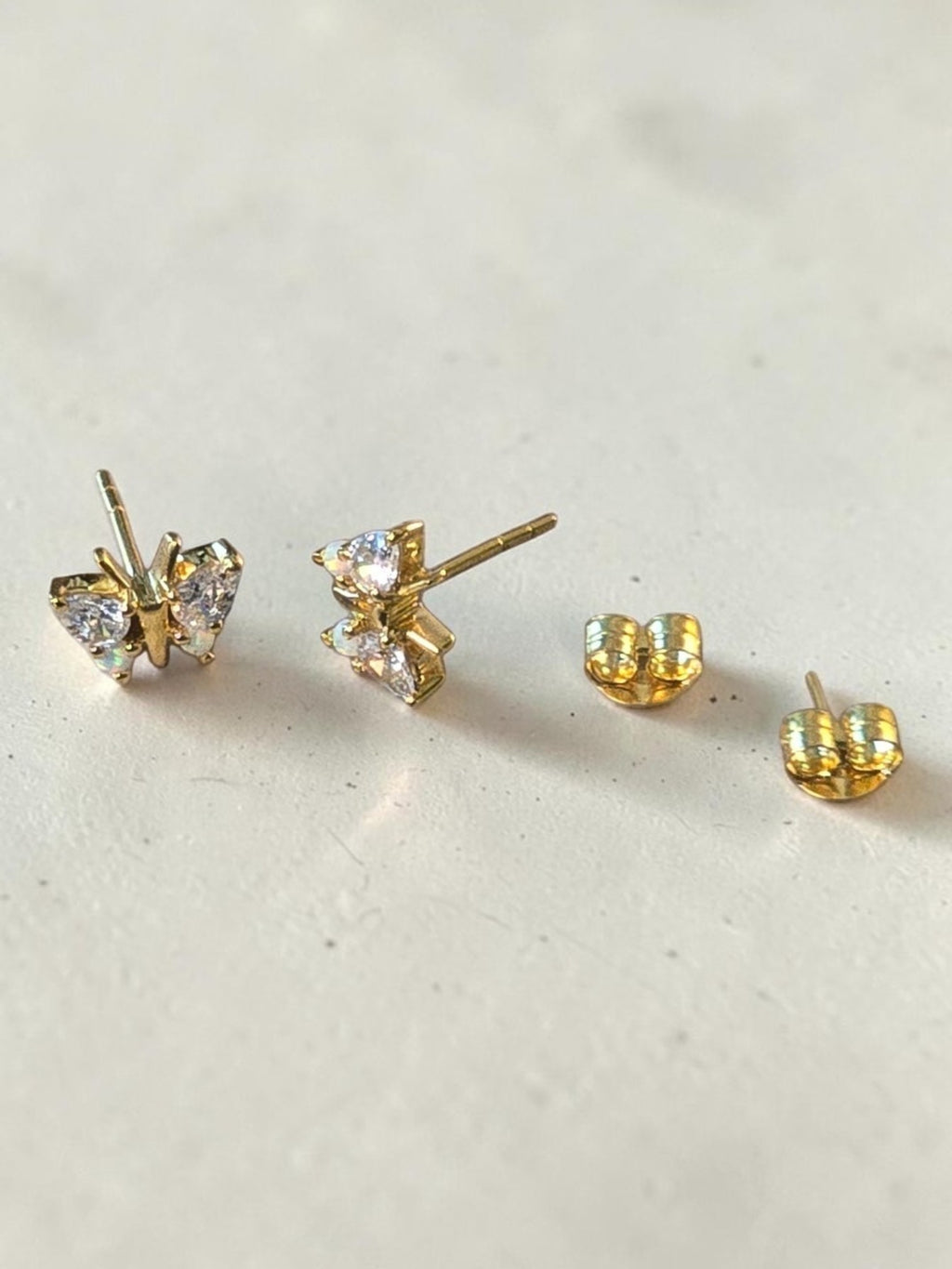 reveriebygemma butterfly stud earrings in 18kt gold vermeil with simulated opal and diamond wings on a textured surface.