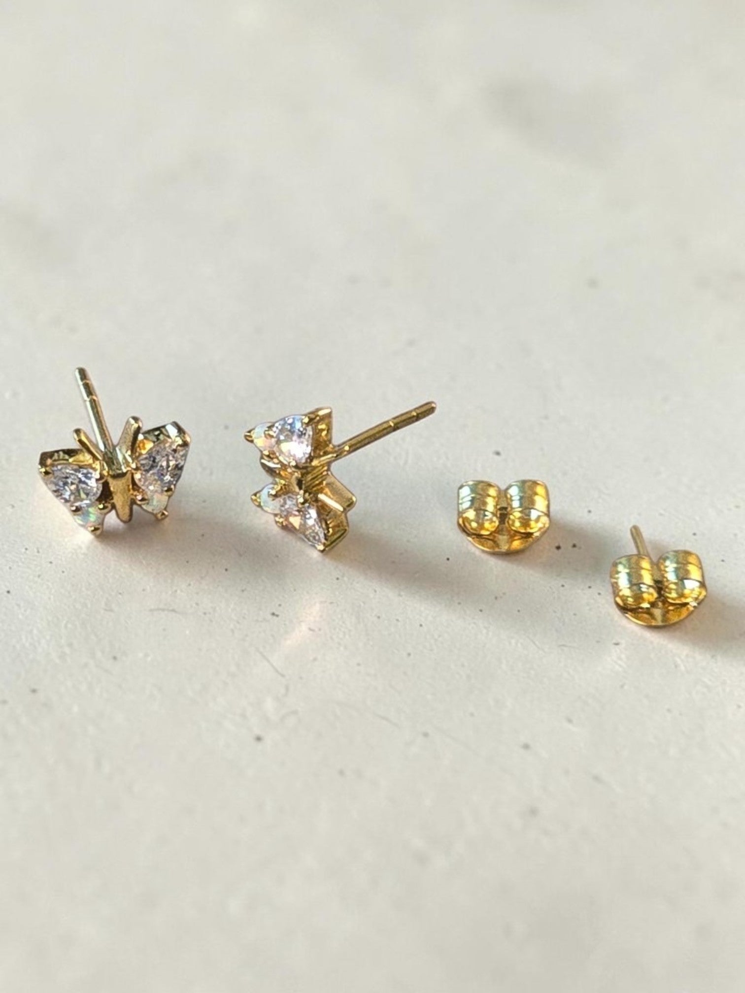 reveriebygemma butterfly stud earrings in 18kt gold vermeil with simulated opal and diamond wings on a textured surface.